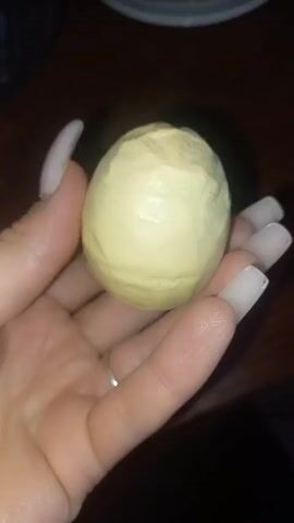 A close-up shot shows a person's left hand holding a light yellow, roughly spherical object. The object has an irregular, textured surface, resembling a small, unpeeled citrus fruit or a ball of dough. The hand has long, light-colored fingernails, some with a light pink or nude polish. A thin silver ring is visible on the ring finger. The background is dark and out of focus, with a hint of a wooden surface. The lighting is dim, casting shadows.