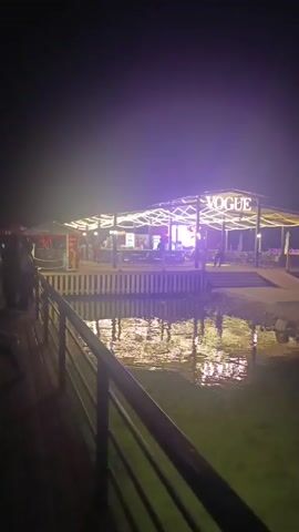 The video opens on a nighttime scene of a restaurant or bar called "VOGUE" illuminated by purple and white lights. The establishment is situated over a body of water, and the lights from the restaurant are reflected on the water's surface. In the foreground, a dark railing runs along a walkway. Several people are visible on the walkway and near the entrance to the restaurant, some appearing to be patrons. The overall atmosphere is one of a lively nighttime gathering place.