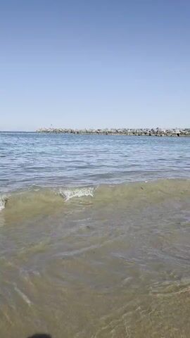 The video captures a serene beach scene with clear blue skies and calm water. In the foreground, gentle waves lap against the sandy shore, with the clear water revealing the sandy bottom. In the distance, a stone jetty stretches out into the ocean, with a small structure, possibly a lighthouse or buoy, at its end. The overall ambiance is one of tranquility and natural beauty.