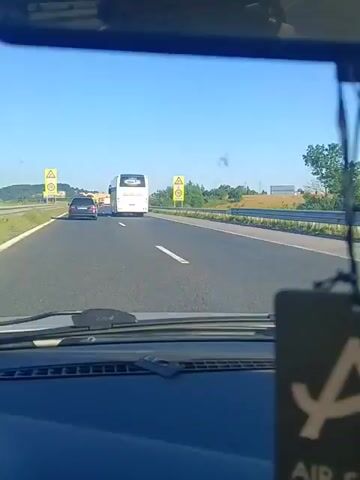 The video captures a view from inside a vehicle driving on a highway. The perspective is from the driver's seat, looking forward through the windshield and also catching glimpses of the road and surroundings in the rearview mirror. In the distance, a white bus is visible traveling in the same direction. A speed limit sign indicating 110 km/h is prominently displayed on the right side of the highway. The highway itself is asphalt with white lane markings. The surrounding environment appears to be rural, with green trees and fields visible beyond a metal guardrail. The sky is clear and blue, suggesting a sunny day. The interior of the car shows the dashboard and the top of the windshield. The video appears to be a continuous shot of the drive.