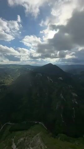 The video shows a mountainous landscape with a winding road. The mountains are covered in dense green forest, and the road snakes through the valleys. The sky is partly cloudy, with the sun occasionally peaking through. The view is from a high vantage point, possibly a mountaintop. There are no people or animals visible in the video.