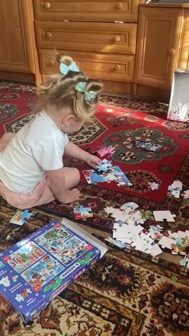 The video shows a young girl with blonde hair in pigtails, adorned with light blue bows, sitting on a red and patterned carpet. She is wearing a white t-shirt and pink shorts. In front of her, scattered across the rug, are numerous pieces of a jigsaw puzzle. The girl is actively engaged in assembling the puzzle, holding a puzzle piece in her right hand and appearing to examine it.

To her left, a blue and white puzzle box is visible. The box features four different puzzle images, some of which depict cartoon characters. There is also a small pink toy horse near the girl. The background consists of wooden cabinetry with drawers, suggesting an indoor setting, likely a home. Sunlight streams in from the right side of the frame, casting shadows on the carpet and indicating it is daytime. The overall atmosphere seems peaceful and focused, with the child engrossed in her activity.

Visible text on the puzzle box includes Russian lettering that appears to be a title and possibly instructions or descriptions related to the puzzle's contents. Some discernible words include "В НАБОРЕ" which translates to "In a set" and "ПАЗЛЫ" which means "puzzles."