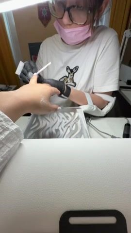 A woman with glasses and a pink face mask is giving a manicure to another person. She wears black gloves and a white t-shirt with a deer print. She holds a white brush and dusts off the client's hand, which is positioned over a white and gray patterned mat. The client's hand has white powder on it. The scene is brightly lit by a lamp overhead.