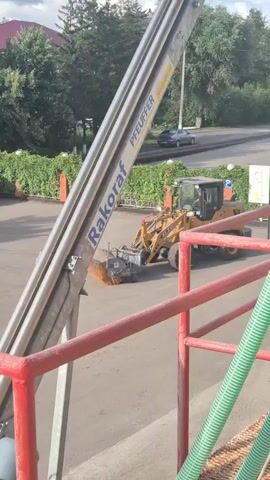 The video shows an outdoor scene in Petropavl City Administration, Kazakhstan. In the foreground, a metal structure with red railings and green hoses is visible. Further back, a yellow loader with a sweeping attachment is operating on an asphalt surface, kicking up dust as it sweeps. The loader appears to be a small construction or maintenance vehicle. Behind the loader, a green hedge forms a barrier, with a fence and trees beyond. Two cars are parked or driving in the background, one silver SUV and another lighter colored sedan. The sky is partly cloudy, suggesting it is daytime with moderate weather conditions. The overall impression is of routine maintenance or cleaning work being carried out in a public or administrative area. There is visible text on a silver, angled object in the mid-ground which reads "Rakoraf PFEUFFER".