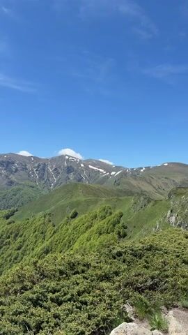 The video shows a wide shot of a mountainous landscape under a blue sky with scattered clouds. The mountains in the foreground are covered with lush green trees and shrubs. Some higher mountain peaks in the distance have patches of snow. The camera pans slowly, showing a panoramic view of the mountain range. The wind seems to be blowing.
