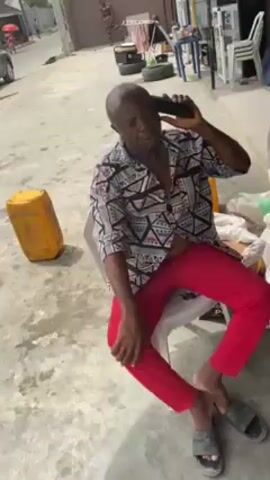 A medium shot depicts a bald man sitting on a white plastic chair outdoors, engaged in a phone conversation. He's wearing a printed short-sleeve shirt and bright red pants, with grey sandals on his feet. A large yellow container sits to his left, and several bags of what appears to be grain or other dry goods are behind him. Two cars, a grey SUV and a white sedan, are visible parked on the street in the background, along with some miscellaneous objects like a small generator and tables and chairs. The man is actively gesturing with his right hand while holding his phone to his ear with his left hand. The pavement is concrete, showing signs of wear and discoloration.