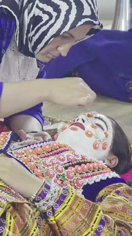 A person wearing a hijab applies makeup to another person lying down. The person lying down is wearing an ornate, colorful garment and has white makeup on their face adorned with small orange and gold decorative elements. Another person, a child, is visible in the background to the right of the person applying makeup. The environment appears to be indoors, possibly a room with a soft surface on the floor.