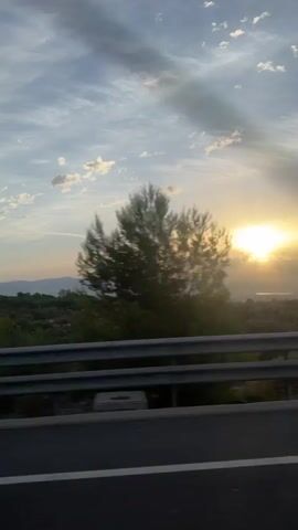 The video shows a view from a vehicle, likely a car, moving along a road. A metal guardrail is visible in the foreground, separating the road from the landscape beyond. The landscape consists of trees and hills in the distance, with the sun setting or rising behind them. The sky is partly cloudy with rays of sunlight breaking through. The overall impression is one of travel through a natural, possibly rural, setting at either dawn or dusk.