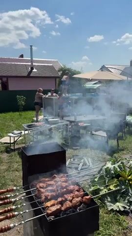 The video shows a backyard scene on a sunny day with a clear blue sky and some clouds. In the foreground, a barbecue grill is smoking, with meat skewers cooking on it. To the left, there is a table with food and grilling equipment. In the middle ground, a large above-ground swimming pool is set up, and several people are in it, including children and adults. Some people are standing around the pool, and one person is on the edge of the pool. A woman in a swimsuit is standing near the pool. There are tables and chairs set up in the yard, and a large umbrella provides shade. In the background, there are houses and trees. The overall atmosphere appears to be a casual gathering with a barbecue and swimming.