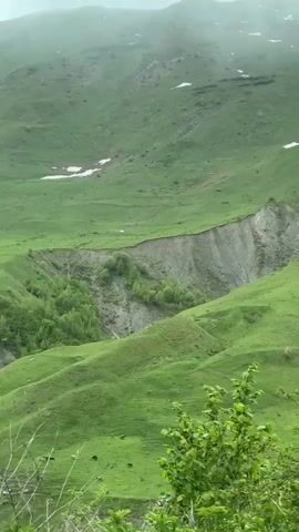 The video shows a lush green mountain landscape under overcast skies. The camera pans slightly from left to right, revealing rolling hills and valleys. There are patches of snow visible on the higher slopes. The dominant color is green, with some brown areas where the earth is exposed, and glimpses of a stream in the distance. The foreground features green foliage, partially obscuring the view.