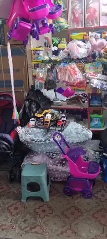 The video shows a cluttered toy store. There are shelves full of toys, including dolls, toy cars, stuffed animals, and other various playthings. In the foreground, there is a pink toy stroller and a small green stool. The lighting is dim and appears to be artificial. The overall impression is one of a densely packed and somewhat disorganized merchandise display.