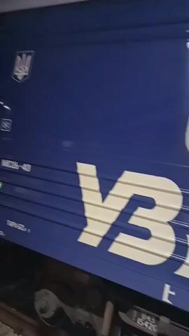 A close-up, angled view of the side of a blue train car. Large white letters that appear to spell "УБ" are visible on the blue surface. To the right, a train window displays illuminated text indicating "ПОТЯГ 52/51" and "ПЕРЕМИШЛЬ-КИЇВ", with numbers "3" and "2" below. A partial view of a train wheel assembly is visible at the bottom of the frame. The Ukrainian trident emblem is visible in the upper left corner.