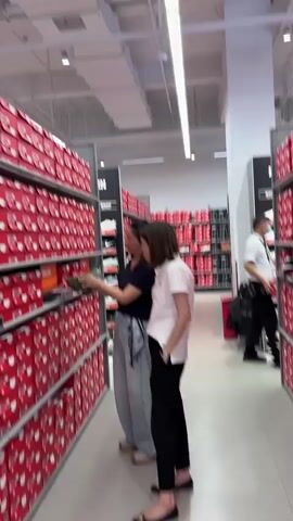 The video shows a wide shot of a retail store's shoe section. In the foreground, on the left, are rows upon rows of red shoeboxes stacked high on shelves. On the right, a black display wall features rows of athletic shoes mounted on individual shelves, each with a price tag. Two women are visible in the middle ground, standing in front of the shoe display. One woman, wearing a dark blue top and gray pants, points at a shoe on the display. The other woman, in a white top and black pants, looks in the same direction. The environment appears to be well-lit with fluorescent ceiling lights. The camera pans slightly, offering a glimpse of more red shoeboxes on the left and the back of the store on the right.
