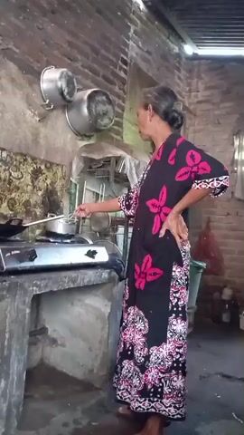 A woman in a black and pink floral dress stands in a kitchen, cooking. She is facing away from the camera, looking towards a stove. She holds a utensil in her right hand, stirring something in a pot on the stove. Her left hand is on her hip. The kitchen appears to be rustic, with brick walls and a concrete countertop. There are pots hanging on the wall and various items on shelves in the background.