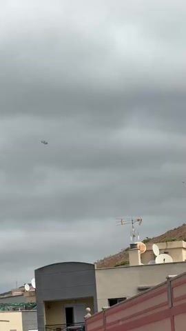 A helicopter flies across a cloudy sky. Below, a rooftop with antennas and satellite dishes is visible. The scene appears to be in a residential area with buildings and a hill in the background.