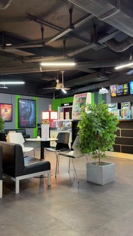 The video shows the interior of a cinema lobby with a seating area and arcade games. The lobby has a black ceiling with exposed ducts and pipes, as well as fluorescent lighting and ceiling fans. The walls are painted in green and gray stripes. There are black and white chairs and tables in the seating area. Several people are sitting at the tables, and some people are playing the arcade games. There are also movie posters on the walls and plants in pots around the lobby. The floor is gray tile.