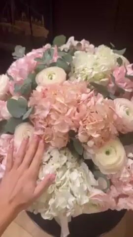The video shows a bouquet of flowers in a white basket. The bouquet is composed of pink and white hydrangeas and cream-colored ranunculus, interspersed with eucalyptus sprigs. A hand touches the flowers gently. The basket sits on a small black side table. The camera pans out, revealing a glimpse of a red chair, a dark brown cabinet, and a mirror. The reflection in the mirror shows a person in workout clothes taking a photo with their phone. There's another bouquet of flowers on the cabinet behind the person in the mirror. The environment appears to be a room in a home or apartment.