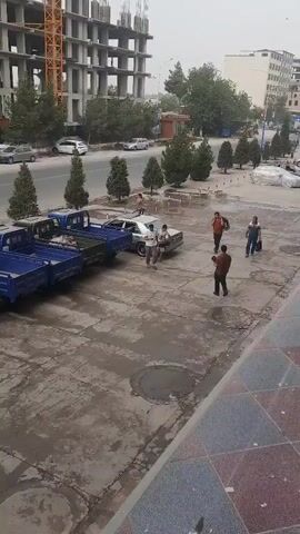 The video shows a street scene with a large unfinished building under construction and a crane in the background. In the foreground, there are several small blue and green trucks parked along the curb. Several people are walking on the sidewalk. A silver car is parked on the side of the street, and a few other cars and a white truck are visible on the road. The overall impression is a busy urban environment with ongoing construction.
