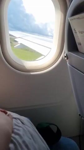 The video shows a view from an airplane window. The wing of the plane is visible outside, as is the ground below which appears to be a grassy field and a runway. The sky is visible with clouds. A person's hand and arm are visible in the lower left corner of the frame. The interior of the airplane, including the window frame and a seat, is also visible. The video appears to be taken during takeoff or landing.