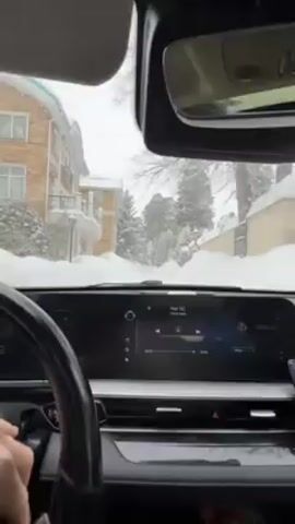 From within a car, the video shows a snowy street. The view is from the driver's seat, focusing on the car's dashboard and the snowy landscape ahead. The dashboard features a large touchscreen displaying music information. Outside, snow falls heavily, blanketing trees and buildings. The camera is fixed, capturing the car's slow movement through the snowy conditions. A hand is briefly visible on the steering wheel.