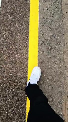 A first-person perspective video shows feet wearing white sneakers walking along a bright yellow line painted on a grey, speckled pavement. The line appears to be a safety marking, possibly at a train station platform or a construction site. The pavement is rough and has small divots. The camera angle is low, focusing on the ground and the walker's legs and feet. The video progresses as the person continues to walk, keeping their feet close to or on the yellow line. The lighting is overcast or dim.