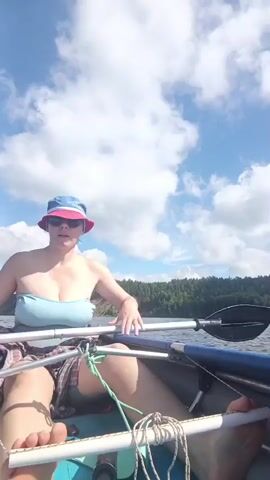A person wearing a blue and red bucket hat and sunglasses is paddling a canoe on a body of water under a cloudy sky. The person is wearing a light blue strapless top and a plaid skirt. The canoe is dark colored and has white ropes and supports. The water is calm and reflects the sky. Trees and a hillside are visible in the background.