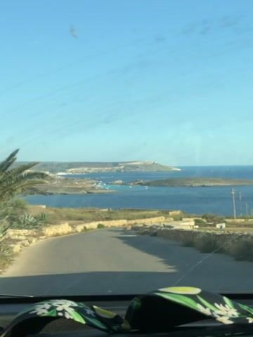 This is a video recording from inside a vehicle, looking out through the windshield. The scene is a scenic coastal road with the Mediterranean Sea visible in the distance. 

The road curves gently to the right as it descends towards the water. On either side of the road, there are low, rough stone walls, typical of rural Maltese landscaping. Lush green vegetation, including palm trees and agave plants, grows along the walls and slopes.

In the background, a wide expanse of the blue sea stretches out to the horizon under a clear, bright blue sky with a few wispy clouds. Several small islands or peninsulas are visible in the bay, with buildings dotting their coastlines. A number of small boats are scattered across the water, suggesting a tranquil day for sailing or leisure. The overall impression is one of a sunny, warm day in a Mediterranean setting.

The time of day appears to be morning or afternoon, given the bright sunlight and clear sky. The location is identified as Qala, Malta. There is no visible text in the video. The view from the vehicle suggests the occupants are on a journey, enjoying the picturesque landscape.