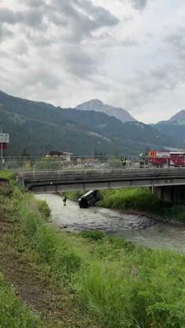 A black car is overturned in a shallow, rocky river. A person in a yellow and black uniform stands in the water near the car, appearing to be a rescuer. On a bridge spanning the river, several people in yellow and black uniforms are observing the scene, and a red fire truck is visible. The background features a densely forested mountain slope and a cloudy sky. The car is tilted at an angle, with its wheels up and its undercarriage exposed to the water. The riverbank on the left is covered in tall green grass and some purple flowers.