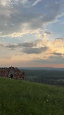 The video opens on a green, grassy hill with a weathered brick archway on the left side. A person stands near the archway, facing away from the camera, looking out over a vast landscape. The sky above is filled with dramatic clouds, illuminated by the soft, warm hues of a sunset. As the video progresses, the camera pans slowly to the right, revealing a wide river winding through a forested valley. The river is dotted with small islands and sandbars, reflecting the colorful sky. The sun is partially obscured by clouds, casting rays of light across the scene. The overall impression is one of natural beauty and serene tranquility.