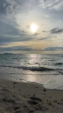 This is a short video clip of a beach scene at Baan Tai, Thailand. The sun is shining brightly through scattered clouds, casting a shimmering reflection on the gentle waves of the ocean. The tide is coming in, with small waves washing over the sandy shore. Footprints are visible in the wet sand, indicating recent human presence. In the distance, a faint outline of landmasses can be seen against the horizon. The overall mood is peaceful and serene, evoking a sense of calm and natural beauty. The time of day appears to be either sunrise or sunset, given the angle of the sun and the soft lighting. The weather is partly cloudy, with the sun breaking through. The location is clearly a tropical beach, with fine sand and clear water. There is no visible text in the video.