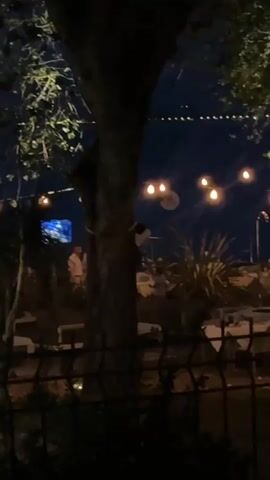 The video shows an outdoor dining area at night. String lights are hung between trees, illuminating tables and chairs set up for patrons. Several people are seated at the tables, appearing to be dining. The overall atmosphere is relaxed and inviting, suggesting a pleasant evening meal.