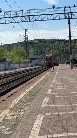 A train is approaching a station on a sunny day. The train is red and gray and has the letters "RZD" on the front. The station platform is made of red and gray bricks. Behind the train is a hill covered in trees. There are power lines overhead.