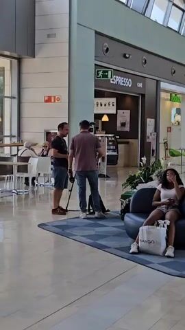 In a well-lit indoor space, possibly a mall or airport, two men are engaged in what appears to be a video shoot. One man, wearing a dark t-shirt and denim shorts, stands to the left, holding a device and seemingly directing or instructing. The second man, in a maroon t-shirt and jeans, operates a video camera mounted on a tripod, facing towards a woman seated on a dark blue sofa. The woman, wearing a white top and shorts, is looking at her phone. A white "MANGO" shopping bag rests beside her on the sofa.

Behind the men, a cafe named "ESPRESSO CAFÉ" is visible, with coffee-making equipment and framed menu items displayed. The overall environment suggests a public, commercial setting, likely during daylight hours given the ample natural light filtering in from large windows. The mood seems professional and focused, with no overt signs of distress or excitement.

The scene unfolds on a blue patterned rug in a spacious, modern interior characterized by tiled flooring and a high, possibly glass, ceiling. Other patrons can be seen in the background, seated at tables in a cafe area. The presence of the camera, tripod, and the interaction between the two men strongly indicate a filming activity. The signage for "ESPRESSO CAFÉ" provides a clear indication of the establishment's purpose. The context of Zaragoza, Spain, places this scene within a specific geographical and cultural setting, though no explicit visual cues in the image directly confirm this location beyond the provided context.