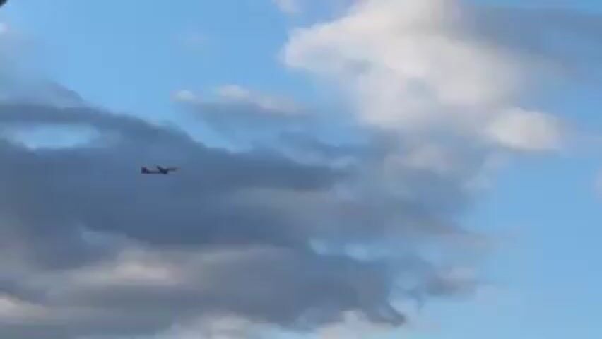 The video shows an airplane flying in a blue sky with scattered clouds. The plane is relatively small in the frame and appears to be distant. The clouds are white and grey. The plane is moving from left to right.