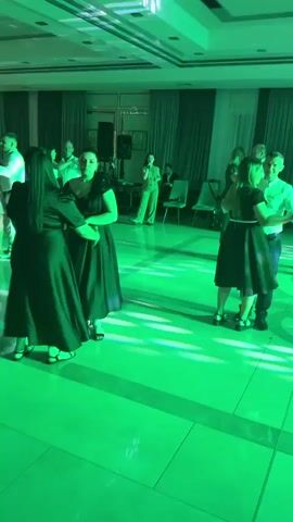 The video shows an indoor event with several people dancing. The lighting changes color, cycling through green and purple. In the foreground, two women in black dresses are dancing together. To their right, a man in a white shirt and blue pants dances with a woman in a black dress. Further back, other couples are dancing, and in the background, a person stands near a speaker and microphone. The room has a polished floor, white walls, and drapes. A chandelier is visible on the ceiling.