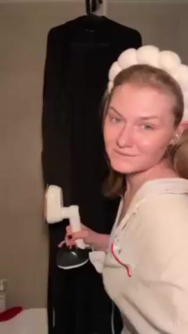 A woman with fair skin and light-brown hair stands in a bathroom, wearing a white shirt and a headband. She holds up her finger as if to emphasize something, then turns toward a black garment hanging on a rack and begins steaming it. She holds the garment open to better access the material with the steamer.