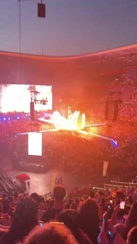 The video shows a large outdoor concert or event in a stadium setting. In the background, a stage is visible with large screens and pyrotechnics creating large flames and smoke. The stadium is filled with a vast crowd of people, many of whom are holding up their phones, some with their lights on, creating a sea of lights. On the stage, a large, golden, humanoid statue-like structure is illuminated and appears to be the focal point of the performance, with lasers projecting from it. The sky above is dark, suggesting it is evening or night, with the stadium lights and stage effects providing the primary illumination. The perspective is from an elevated seating area within the stadium, looking down and across the stage and crowd.