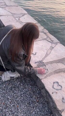 The video opens with an overhead shot of a young woman kneeling on a stone walkway next to a body of water. She has long, straight, reddish-brown hair and is wearing a dark-colored jacket and dark pants. Her left hand is actively arranging small, grey, smooth pebbles on the light-colored stone surface. To her left, the letters "S O S" are clearly formed from these pebbles. To her right, a heart shape made of similar pebbles is already complete. The water beside her is calm and reflects the soft light of the sky.

As the video progresses, the camera slowly pans upwards, revealing a breathtaking sunset or sunrise scene. The woman remains in the lower left of the frame, continuing her activity. The expansive body of water, likely a sea or a large lake, stretches into the distance. Across the water, rolling hills or mountains are visible, silhouetted against a sky painted with delicate hues of pink, orange, and light blue. Lights twinkle on the distant shore, suggesting a town or village nestled at the foot of the hills. A small, dark buoy can be seen floating in the middle distance of the water. The overall impression is one of tranquility and natural beauty, with the soft, diffused light of dusk or dawn illuminating the scene.