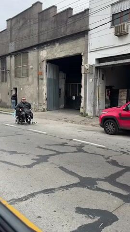 A person in a motorized wheelchair with prosthetic legs crosses a street. The person is wearing a black jacket and dark pants and is carrying a blue and white bag. A red car and a white van are visible in the background. The street is paved and appears to be in an urban area. The weather is overcast. The video is captured from the perspective of a car.