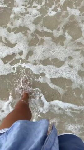 The video shows a first-person perspective of someone walking along the shore of a beach. The camera is angled downwards, focusing on the individual's legs and feet as they step into the shallow water and onto the wet sand. The water is clear and foamy, with small waves lapping at the shore. The sand is light in color and appears to be composed of fine grains with occasional small shells or debris. The person is wearing blue shorts and has a tattoo of a butterfly on their left thigh, as well as a white anklet on their right ankle. The overall impression is one of a peaceful and leisurely walk by the sea.