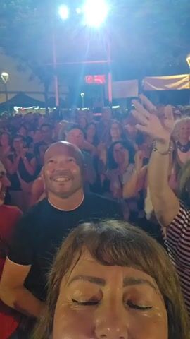 A dark-haired woman records a selfie video, revealing a crowd of people in the background, mostly smiling and looking towards a brightly lit stage. A bald man in a black shirt stands directly behind the woman, also looking towards the stage. The crowd is diverse in age, with many clapping or raising their hands. The environment is an outdoor nighttime event, with trees and tents visible in the background.
