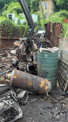 The video shows a scrap metal yard. A large mechanical claw is visible at the top left of the frame, attached to a crane. The claw is grasping a pile of metal debris. In the foreground, a large, rusty, cylindrical object, possibly a muffler or tank, lies on the ground. To its right is a green metal barrel. Other metal objects, including engine parts, pipes, and corrugated metal sheets, are scattered throughout the scene. Vegetation is growing in the background, suggesting an outdoor environment. The ground is muddy and covered in debris. The overall impression is one of disarray and industrial waste.