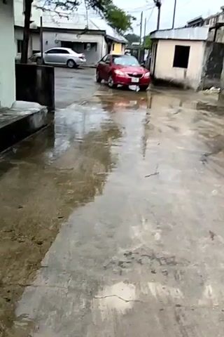 The video shows a residential street after a rain. The ground is wet and reflective. There is a red car parked on the right side of the road. In the background, a silver car is parked near a building. There are other buildings and a tree visible. The sky is overcast. The camera pans slightly to the left. There are no people visible.