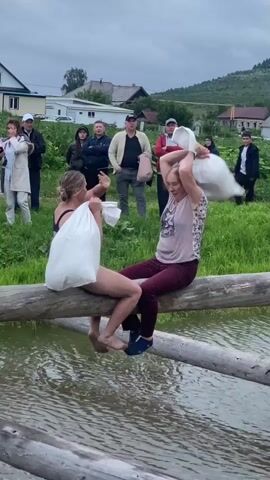 Two women are sitting on a log bridge over water, each holding a large white sack. The woman on the left is wearing a dark bra and underwear, and is partially submerged in the water. The woman on the right is wearing a patterned shirt and burgundy pants. Both women are facing each other and appear to be in a playful or competitive stance. Several people are standing on the grassy bank behind them, watching. The environment appears to be rural with houses and hills in the background. The sky is cloudy.