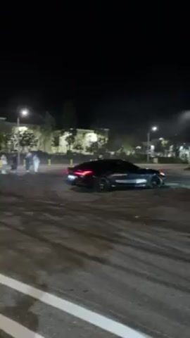 At night, a black coupe is performing a donut, screeching its tires and creating white smoke.  There are several people standing around in a parking lot, appearing to watch the car. Some have cell phones in their hands, as if recording the car.  Street lights illuminate the scene. There are tire marks on the asphalt. Buildings and trees line the background.