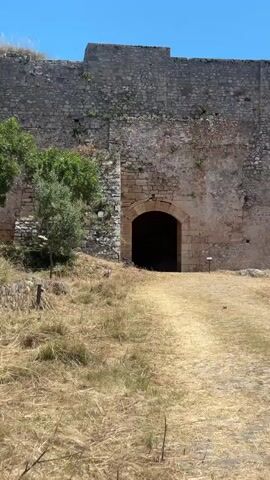 The video shows an arched entrance of a stone wall, surrounded by dry, grassy terrain. There are a few green trees nearby, and a low stone wall in the distance. The sky is bright blue with a few clouds. The video pans slightly from left to right, showing more of the surrounding area.