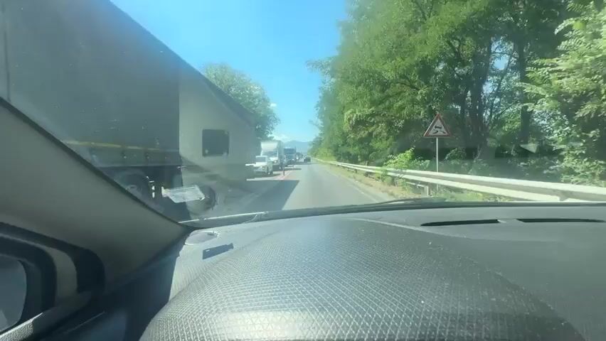 The video shows a first-person perspective from inside a car, looking forward through the windshield. The car is in traffic on a sunny day. Multiple vehicles are visible on the road ahead, including trucks and cars. Trees and foliage line both sides of the road, with a guardrail visible on the right. The sky is clear and blue. The dashboard of the car is in the foreground.