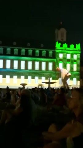 A nighttime outdoor event features a building with a facade illuminated by green and yellow lights. A projection on the building appears to depict a figure. A crowd of people is seated in the foreground, facing the building. The atmosphere is dark, suggesting it is nighttime.