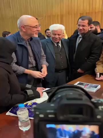 A group of people are gathered around a table in a room with wood paneling. There are two men in suits, one with white hair and the other with dark hair and glasses, standing next to a bald man in a vest and glasses who is signing a book. A woman in a hijab is also standing at the table. There is a camera pointing towards the group, and a book and water bottle on the table. The people are smiling and appear to be in a friendly and informal setting.