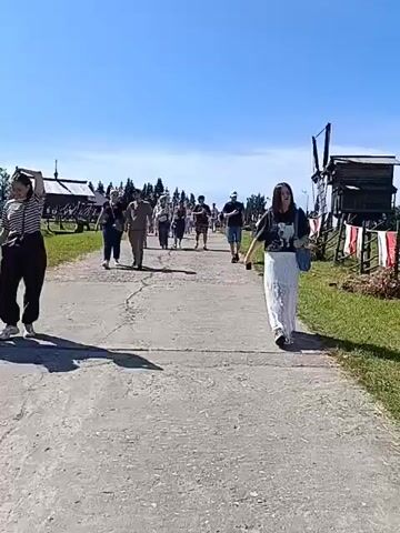 A group of people walk down a paved path on a sunny day. The path is bordered by grassy areas and some wooden structures on either side. In the background, there are trees and a clear blue sky. The people are casually dressed and appear to be walking in the same direction. The camera is positioned at a low angle, looking forward along the path.